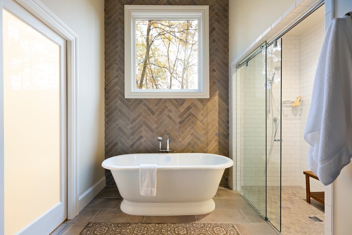 Free-standing tub with herringbone tile feature wall in a bathroom remodel in Charlotte, NC by Distinctive Design + Build + Remodel.